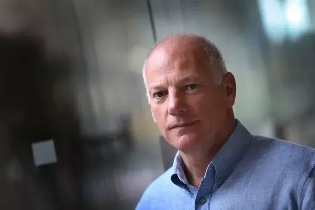 A bald man in a blue shirt is standing in front of a glass wall and looking at the camera.