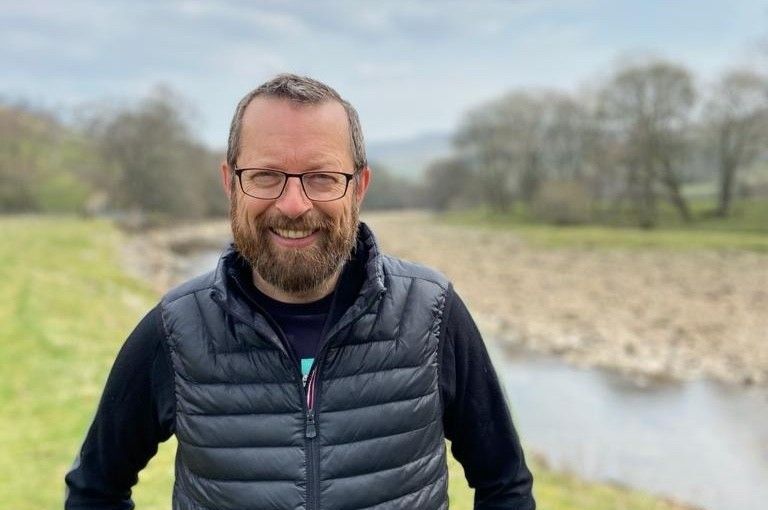 A man with a beard and glasses is standing in front of a river.