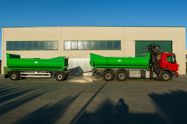 Un camion con cassone ribaltabile verde con rimorchio parcheggiato davanti a un edificio beige in una giornata di sole.