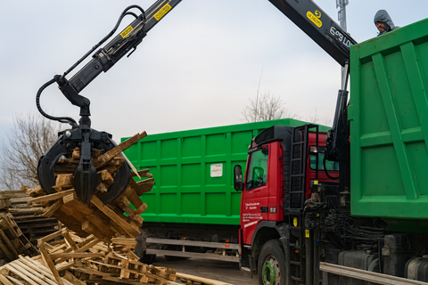Una gru carica pallet di legno su un camion verde, è visibile la cabina di un camion rosso.