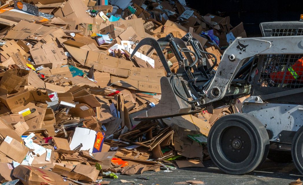 Una pala compatta con attacco a pinza, che raccoglie cartone e rifiuti riciclabili in un grande mucchio.