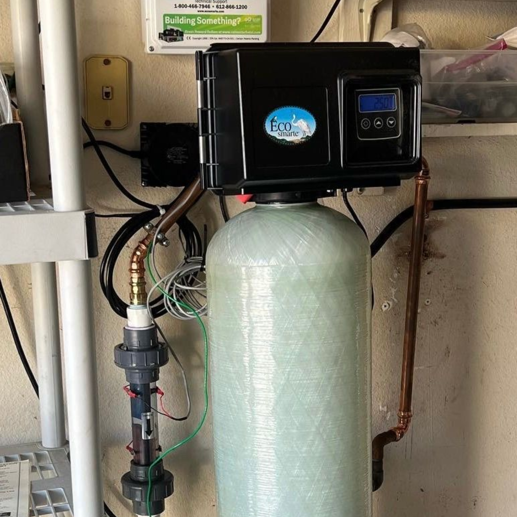 Water filtration system with black control panel, tall tank, and connected pipes against a wall.