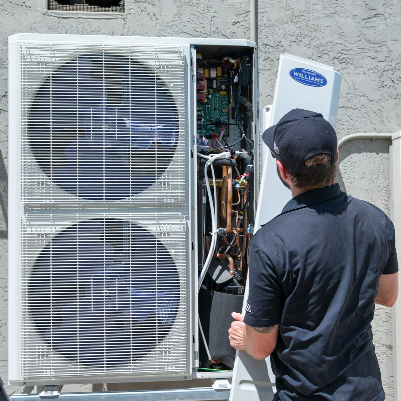 Man removing a panel from an air conditioning unit. Outdoors, sunny. Unit is white, man wears black cap and shirt.
