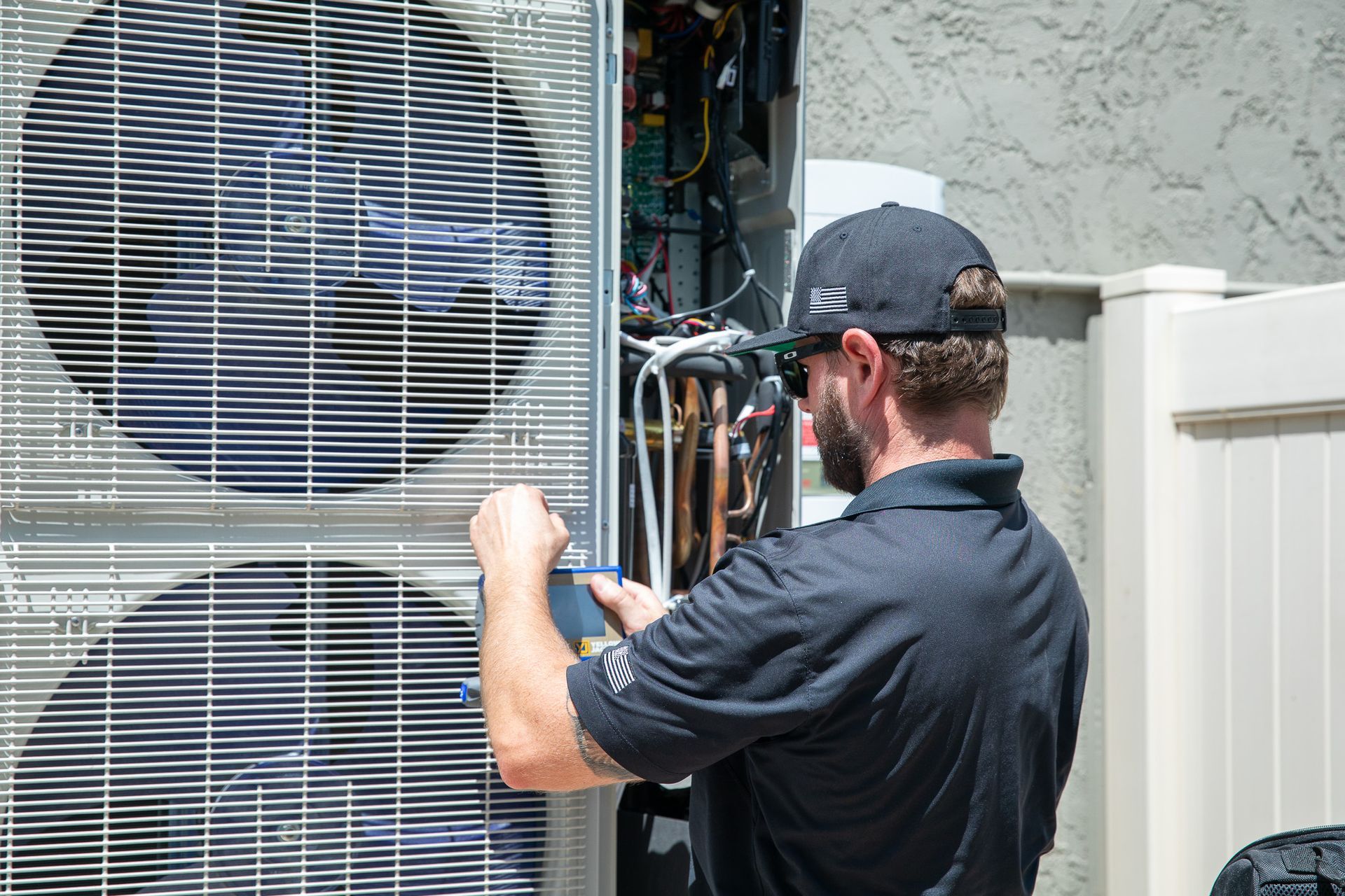 HVAC technician in black shirt and hat, inspecting outdoor AC unit, using a tool.