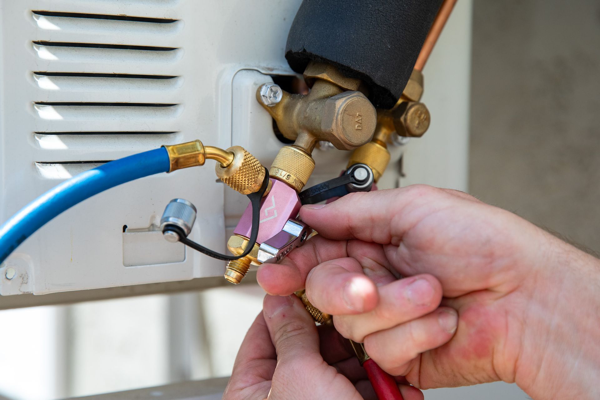 Hands connecting gauges to an air conditioning unit's service valve. Blue, black, and red hoses are attached.