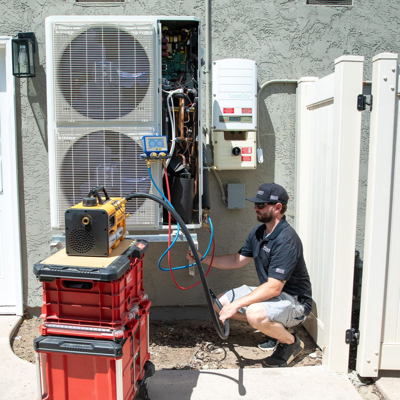 HVAC technician repairs an outdoor unit; using tools. He kneels next to the equipment. Outdoors, near a fence.