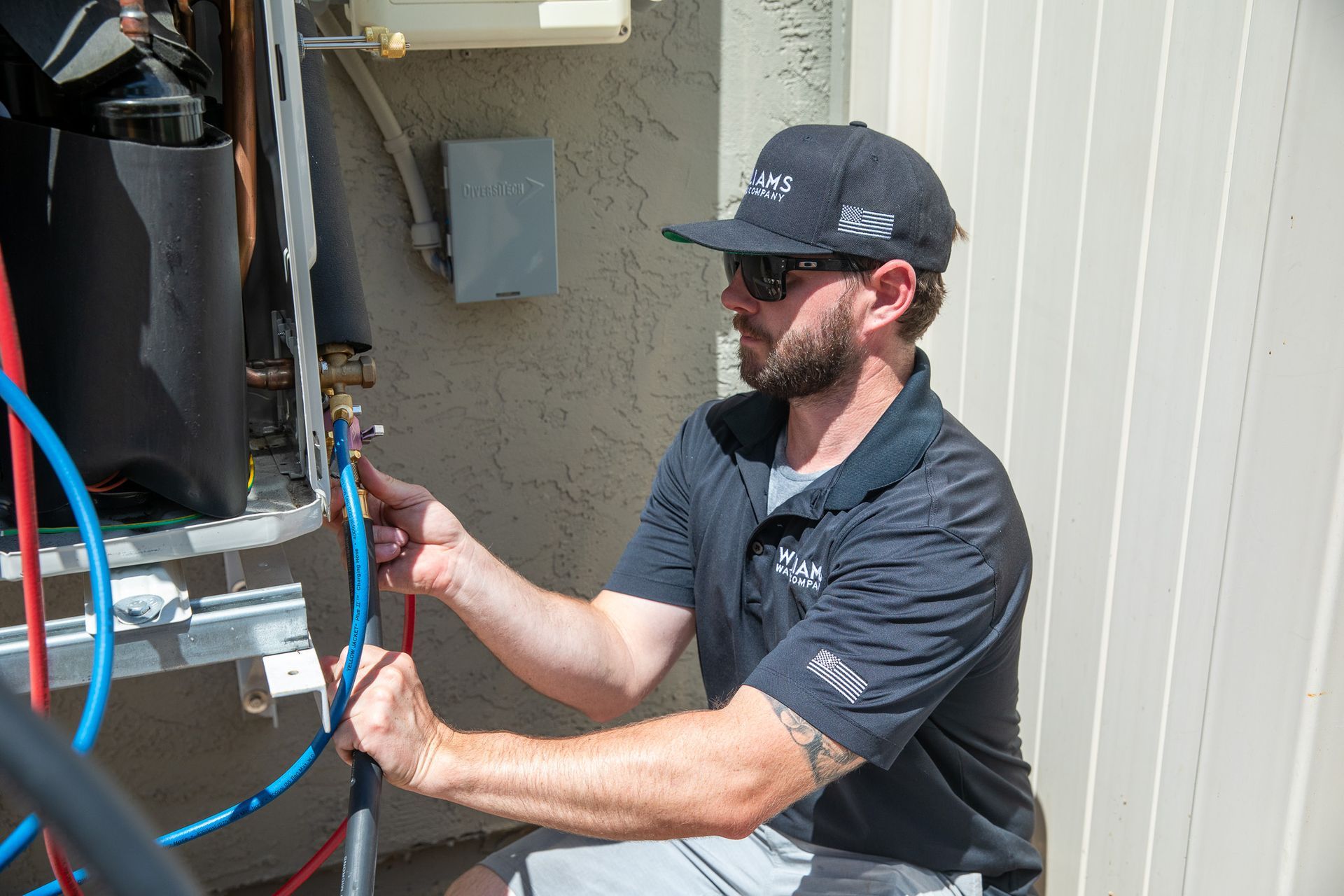 HVAC technician connecting hoses to a unit outdoors. He wears a hat and sunglasses.