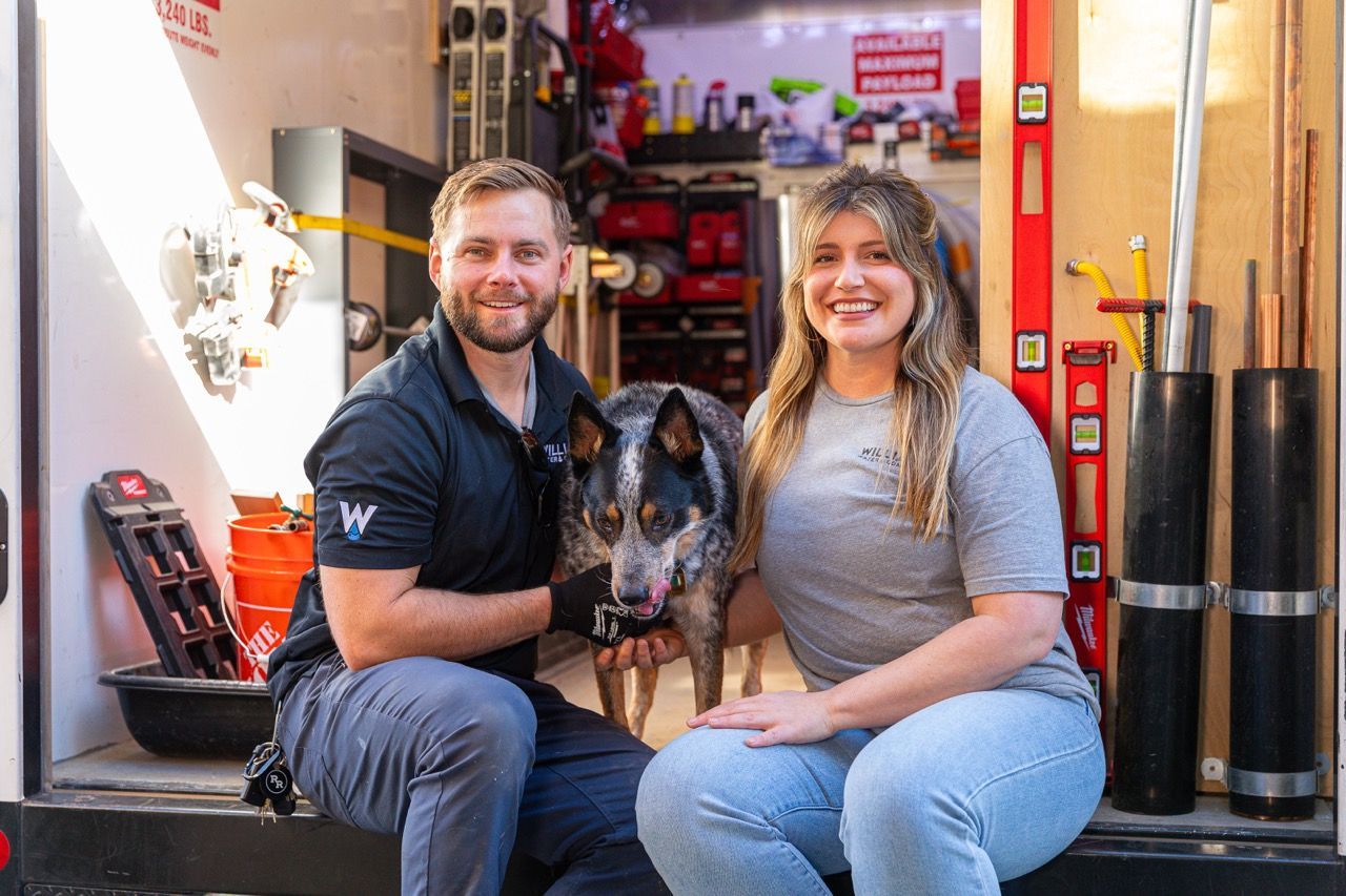 Man, woman, and dog smile in front of a work trailer. Tools and supplies are visible in the background.