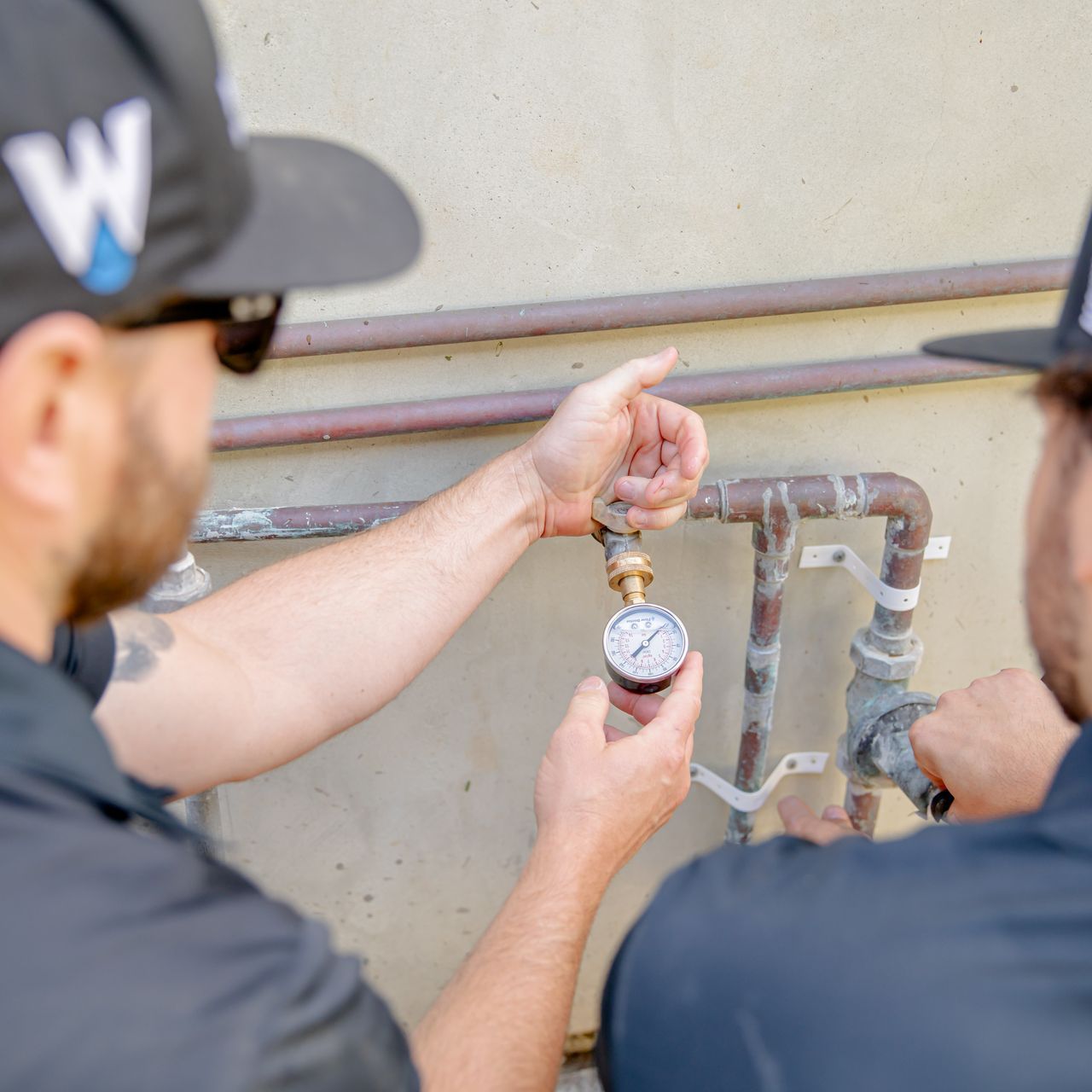 Two men are working on a pipe and one of them is wearing a hat with the letter w on it.