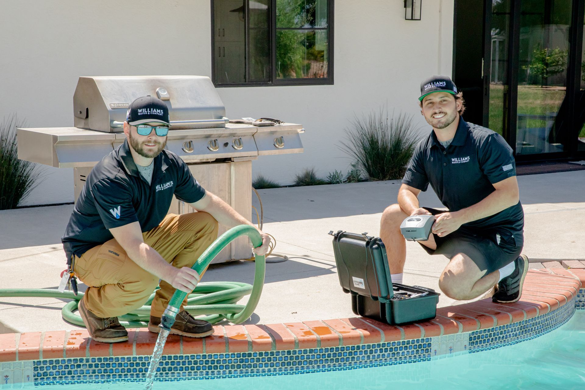 Two men by a pool: one with hose, one with equipment. Both in black shirts and caps.