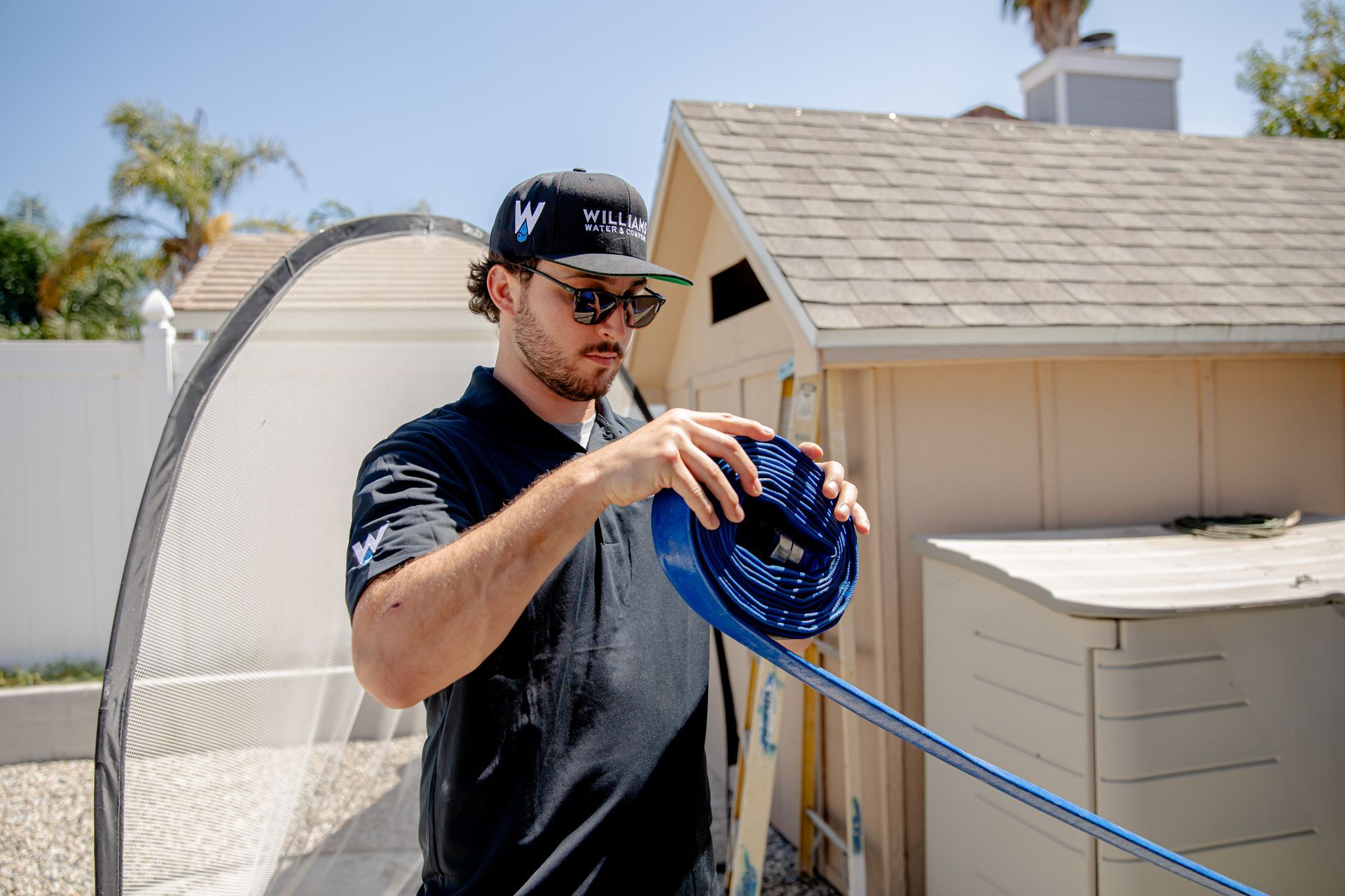 Man with a blue hose preparing to work in a backyard. He is wearing a hat, sunglasses, and black shirt.