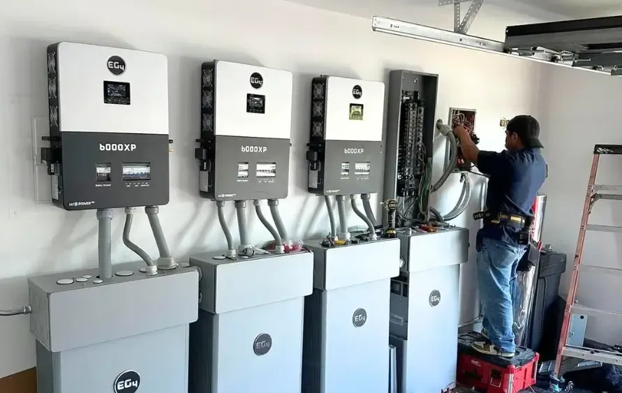 Electrician installing solar energy equipment in a garage. Inverters and batteries are visible.