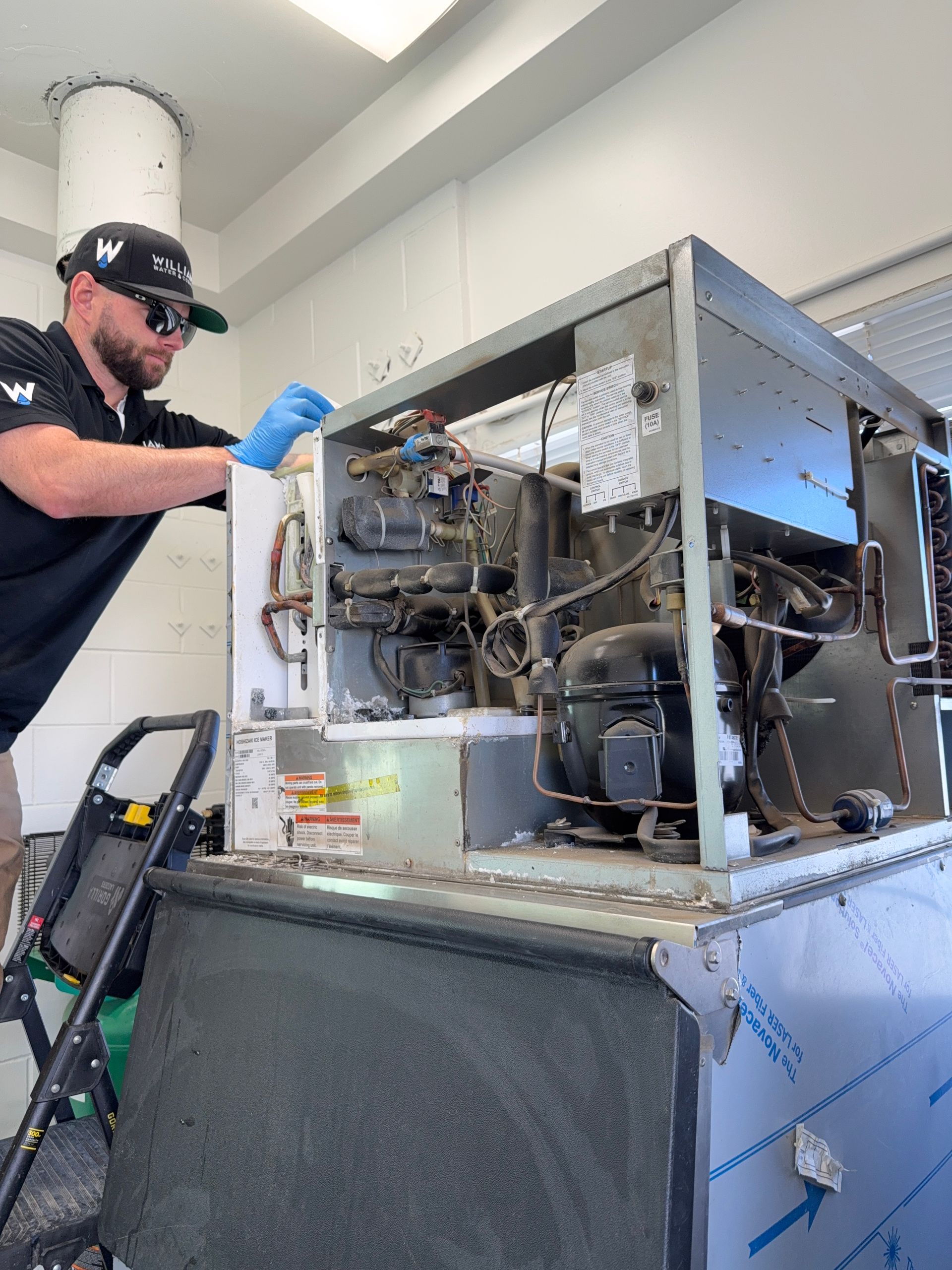 A technician in a cap and blue gloves inspects the internal components of a large stainless steel ice machine.
