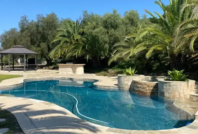 A freeform blue swimming pool with stone coping, a gazebo, and palm trees in a lush garden.