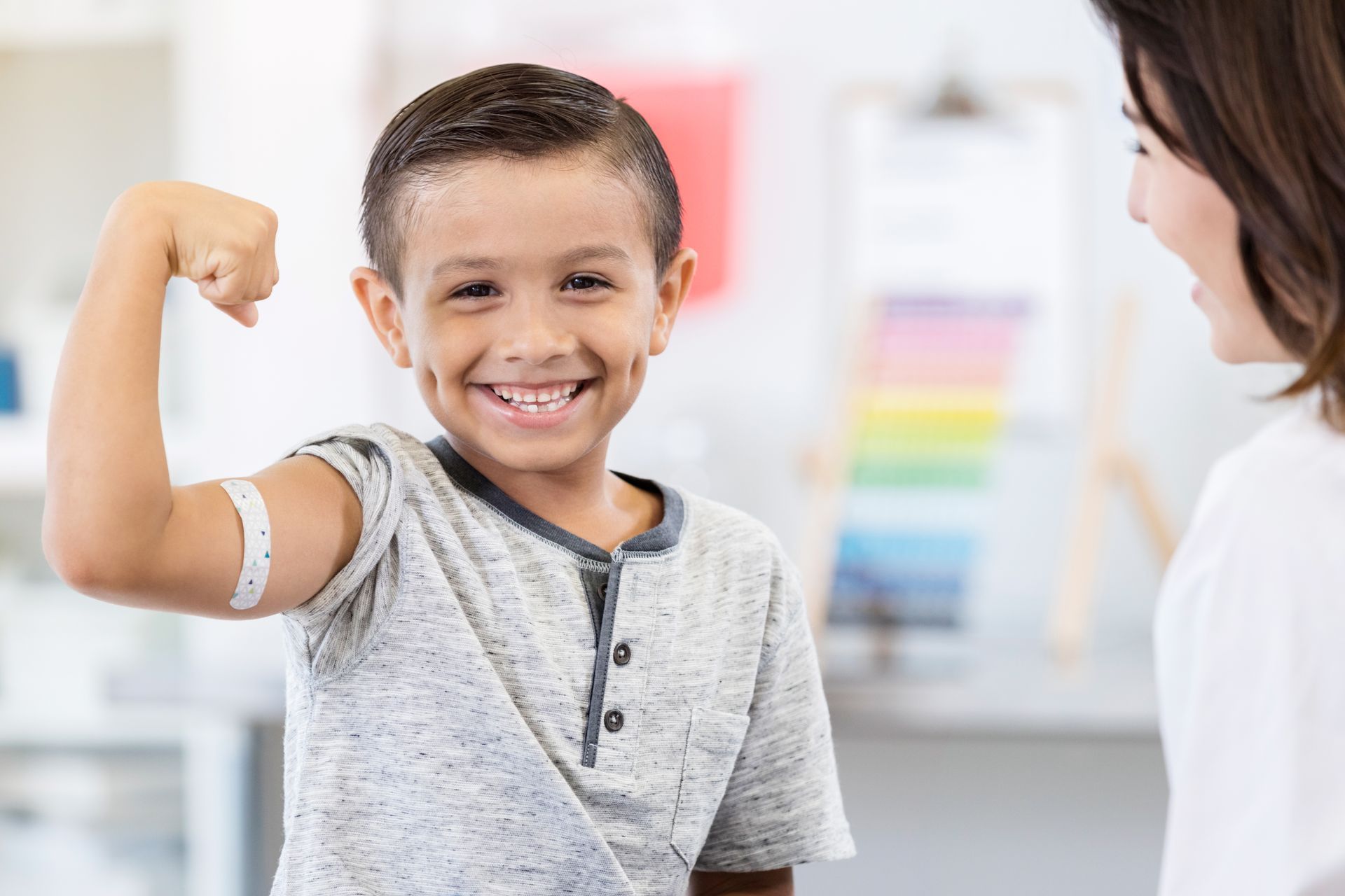 A smiling boy is flexing his arm to show a bandage from a vaccine