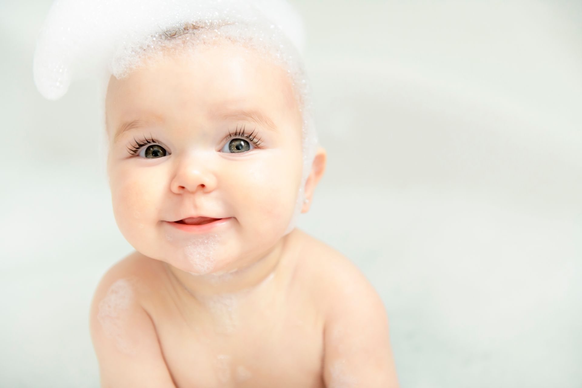 Baby in a bathtub, covered in bubbles, smiling.