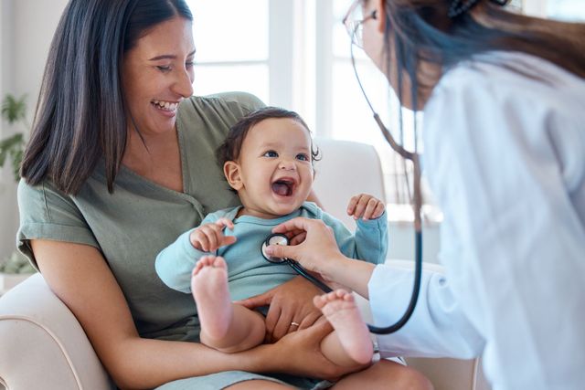 Woman holding smiling baby while doctor examines them in a bright clinic room