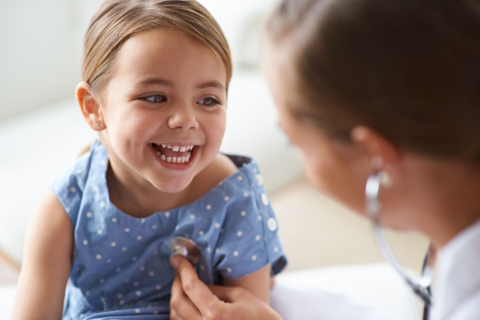 A young girl with pigtails smiles while a doctor listens to her heart with a stethoscope.