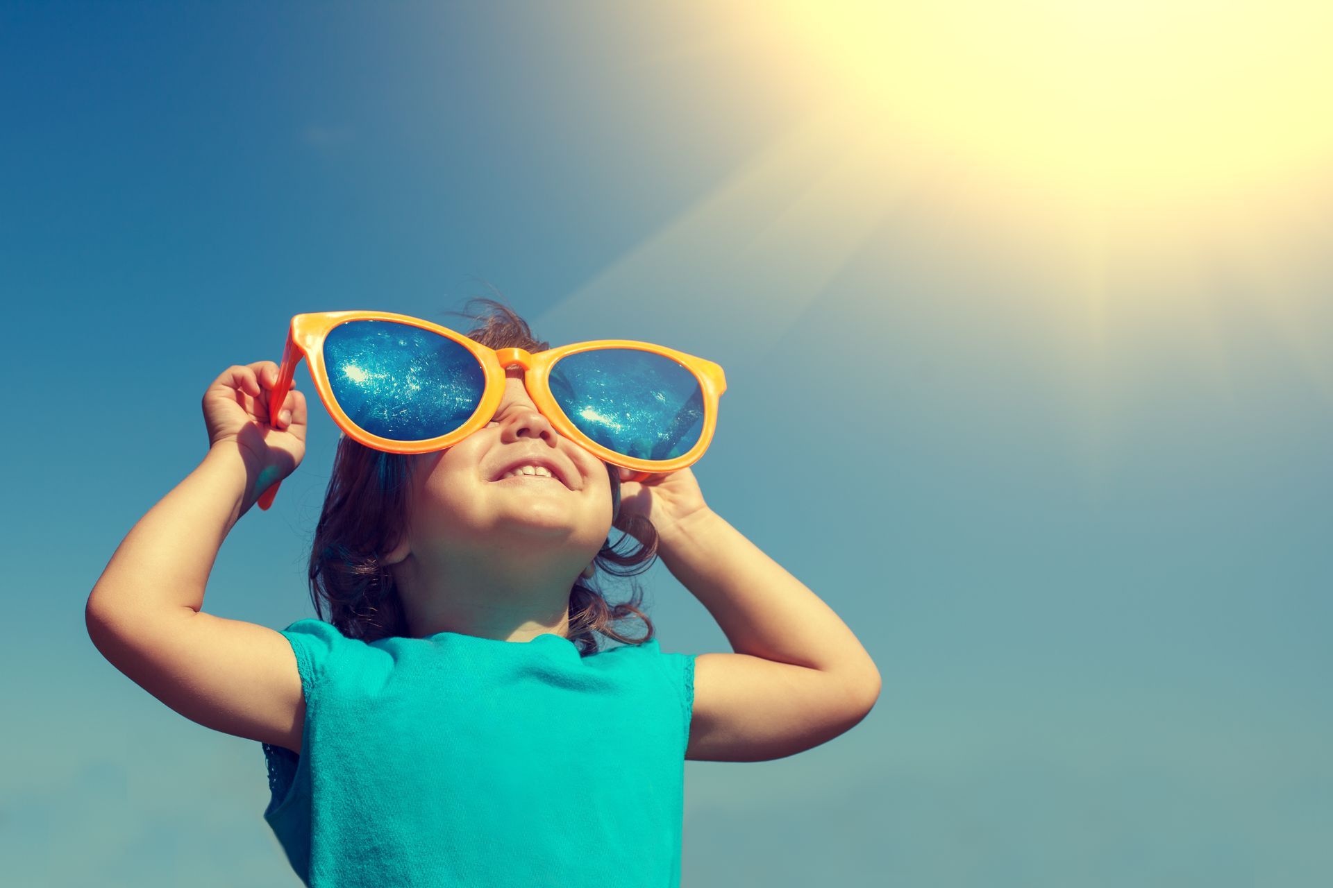 A child with oversized sunglasses is looking up at the sun