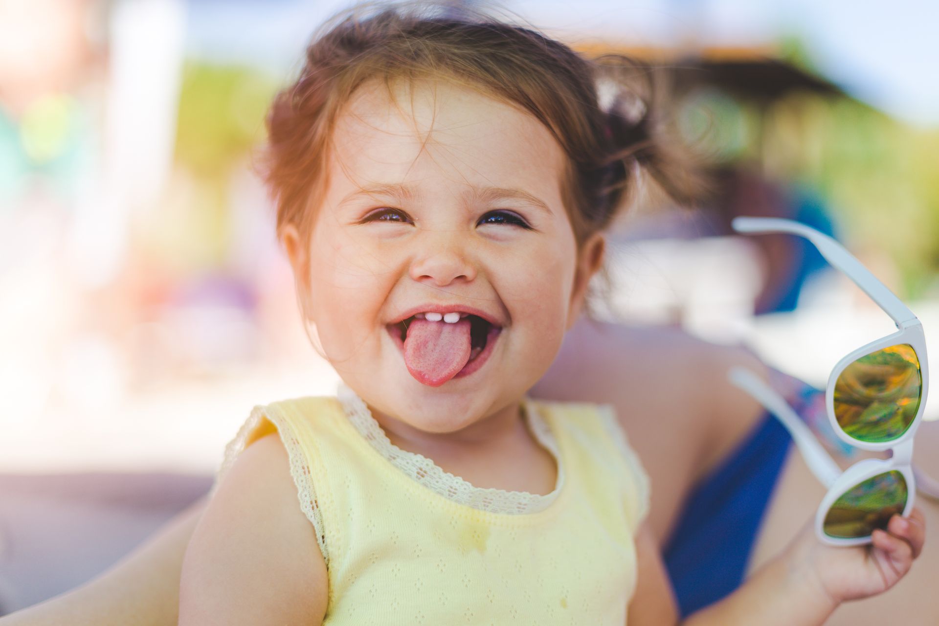 Smiling toddler with tongue out holding sunglasses outdoors on a sunny day
