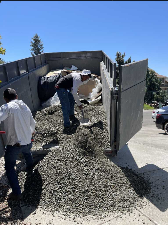 Two men are working on a pile of gravel in front of a dumpster