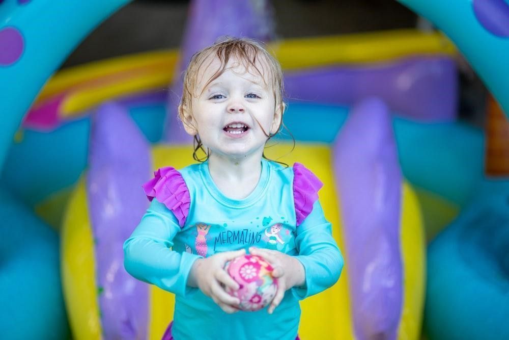 A little girl is holding a ball in front of a bouncy house.