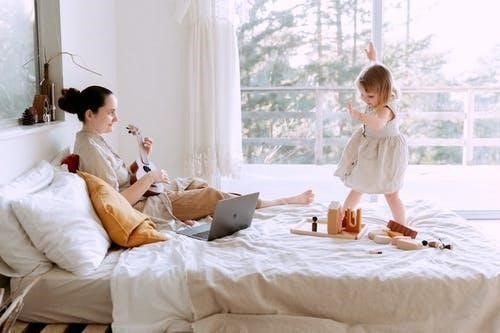 A woman is sitting on a bed with a laptop and a little girl is playing on the bed.