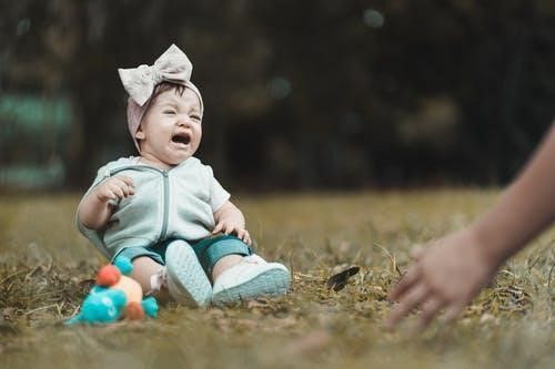 A baby is crying while sitting on the grass next to a toy.