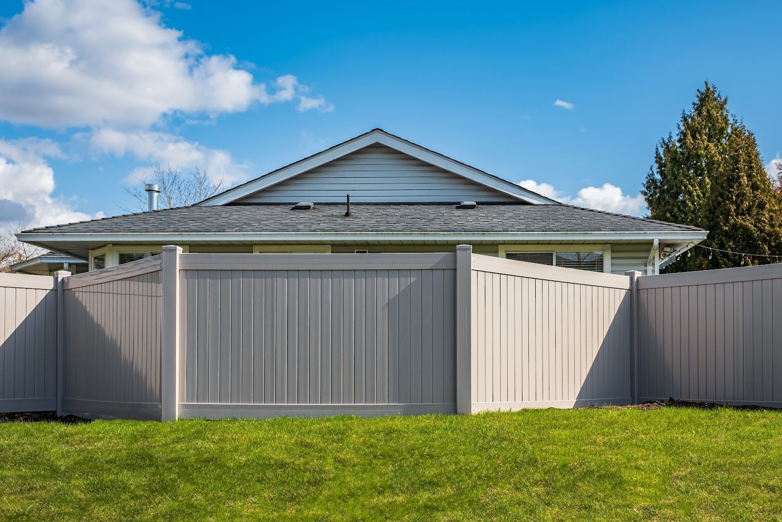 Gray privacy fence in front of a house with a gray roof, on a green lawn, under a blue sky.