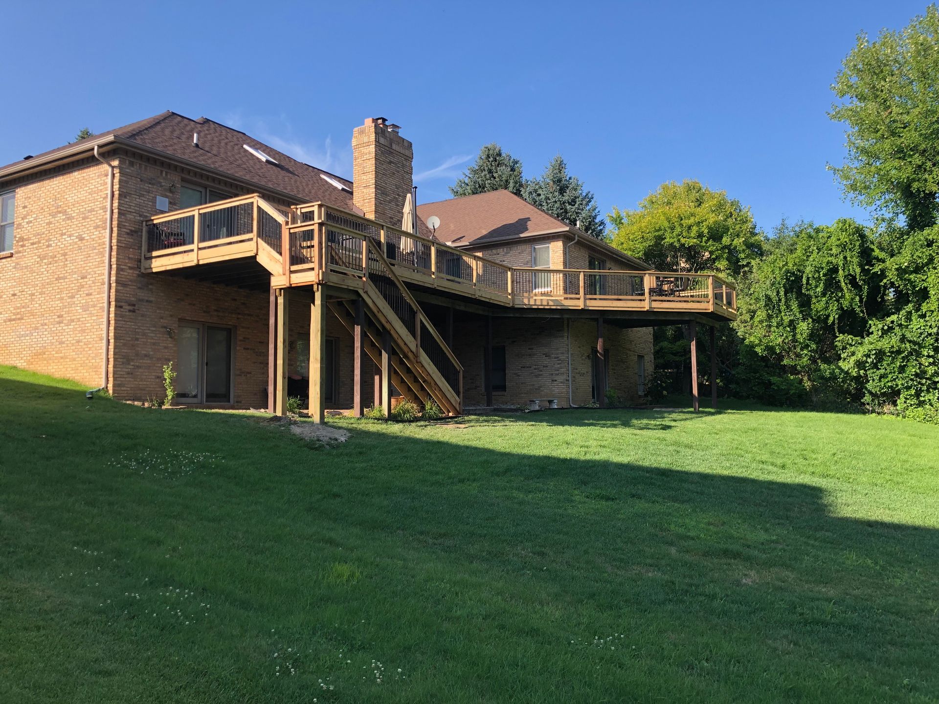 Large wooden deck attached to a brick house, overlooking a grassy yard under a blue sky.