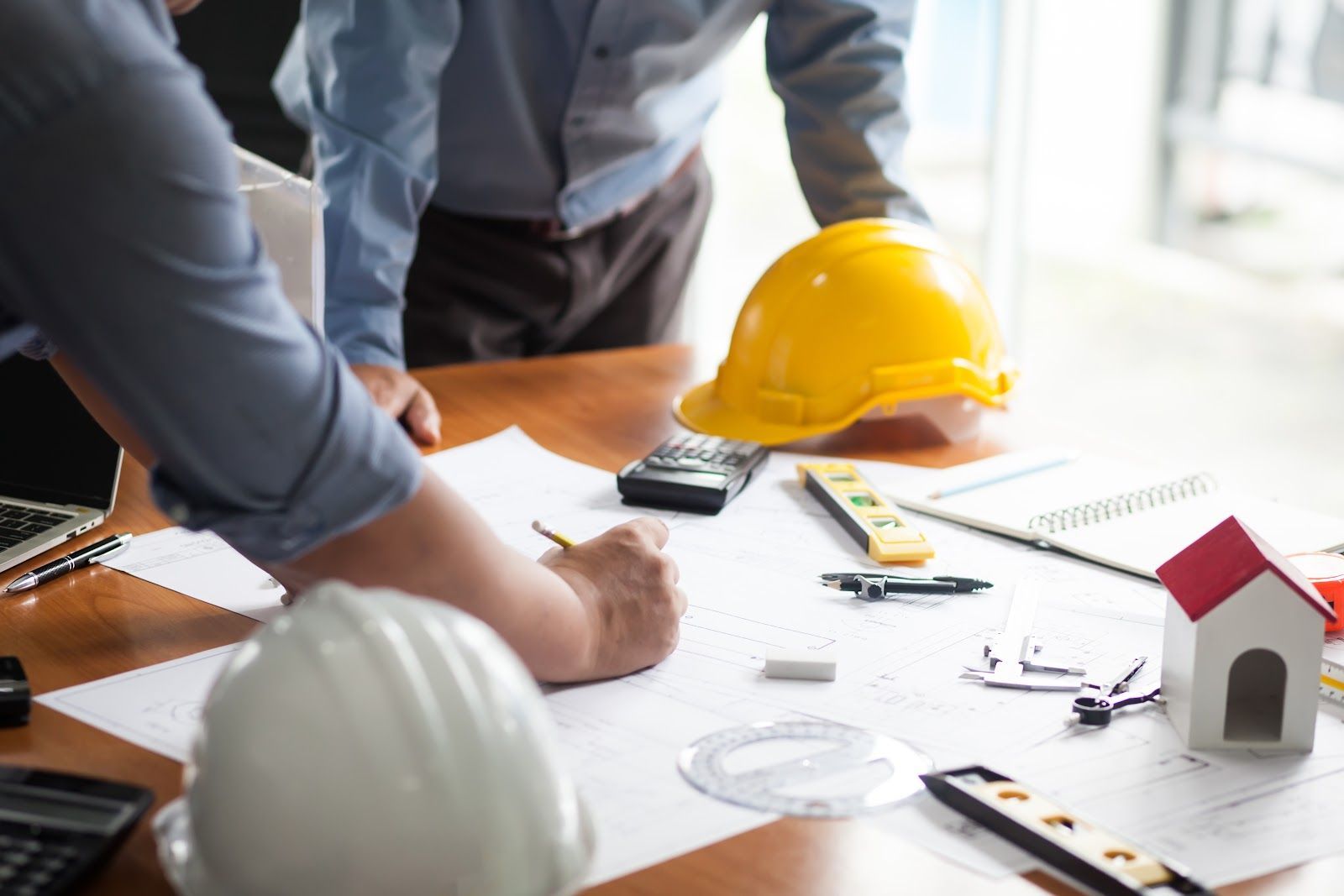 Two people reviewing blueprints at a table with a yellow hard hat and small house model.