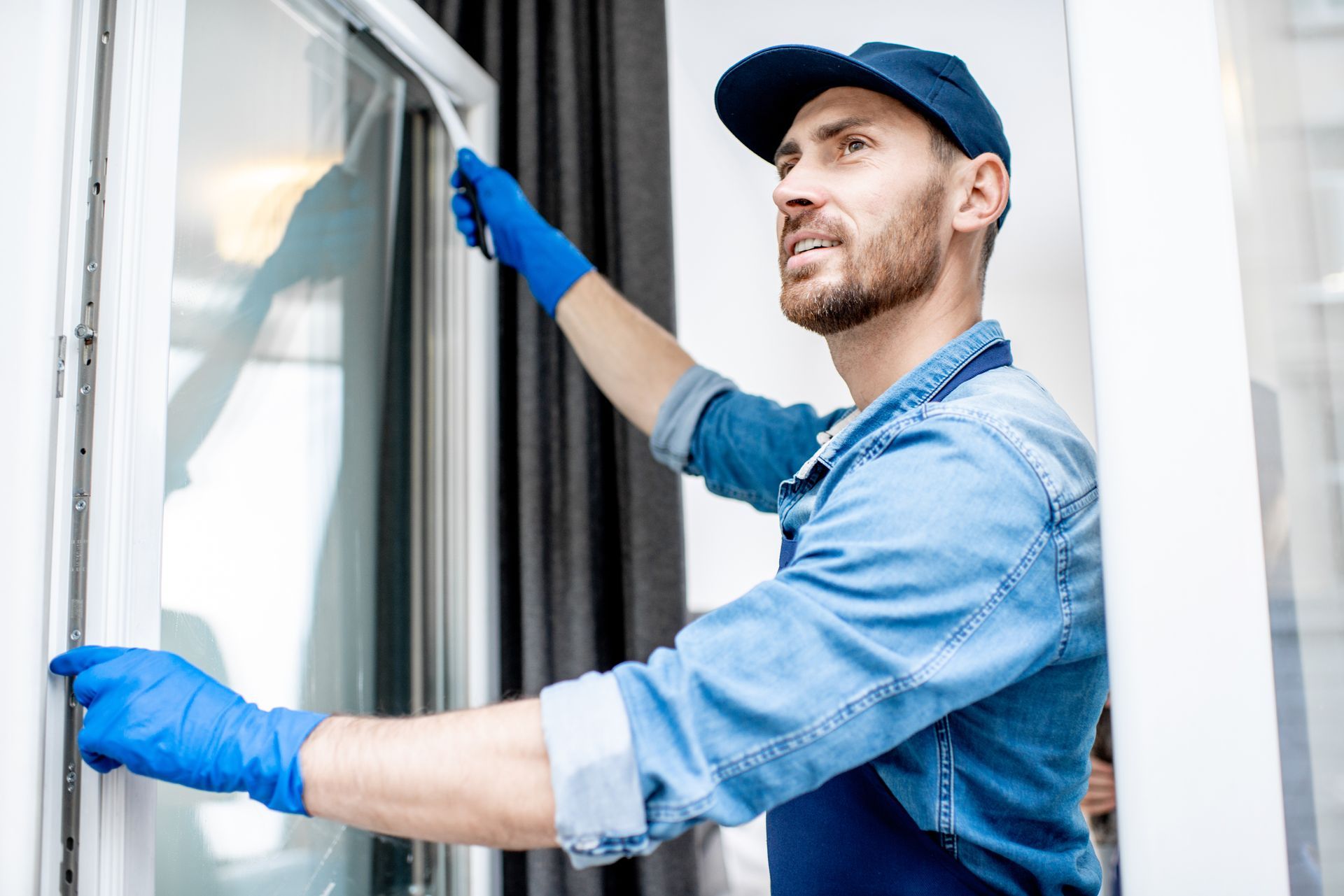 Man in blue gloves cleaning a window, wearing a cap and denim shirt, inside a well-lit room.