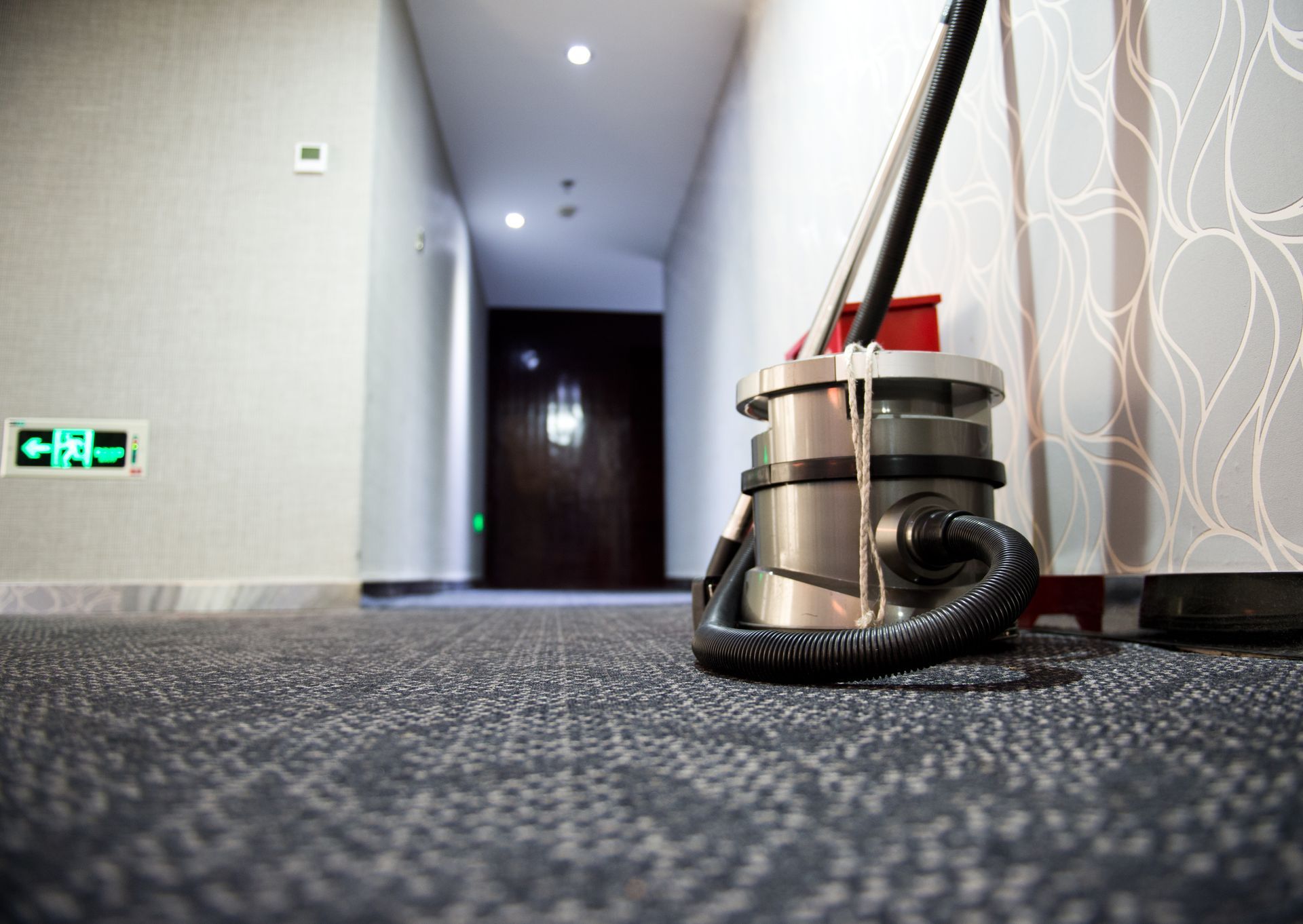 Close-up of a vacuum cleaner, as it stands on top of the carpeted floor in a hotel’s corridor.