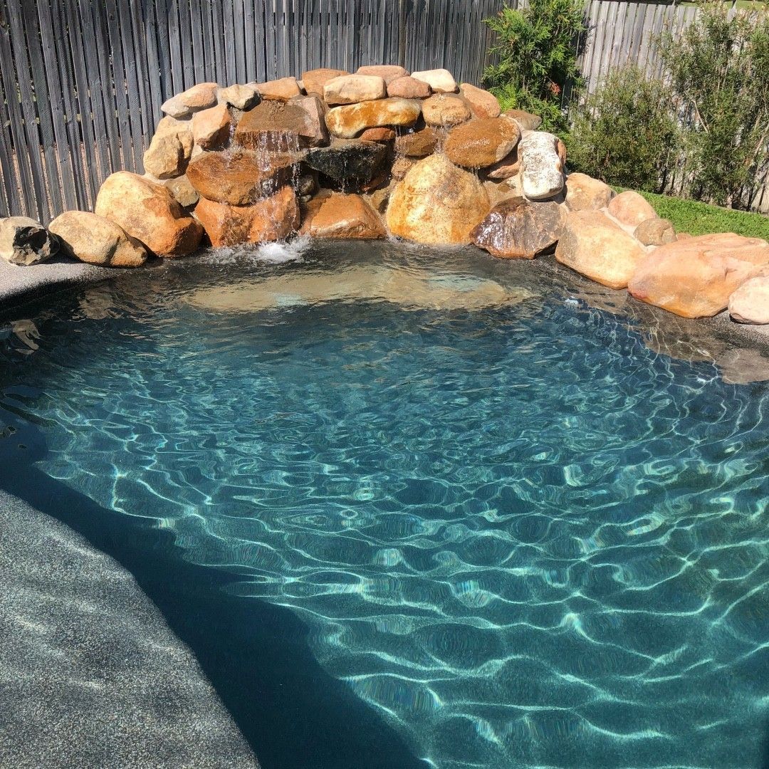 Pool with a rock waterfall. Clear blue water reflects sunlight. Stone and wood fence in background — Tru Blu Pools & Spas in Bluewater, QLD
