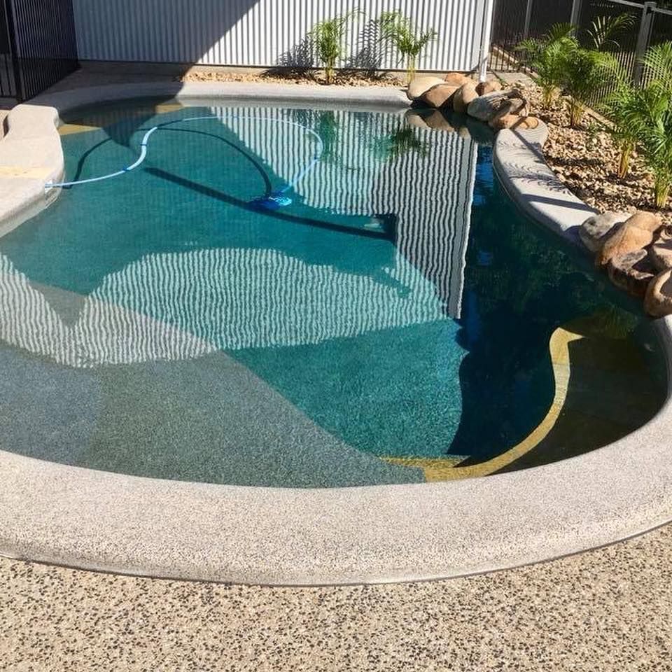 A turquoise swimming pool with a curved edge and a concrete surround. A pool vacuum rests in the water.— Tru Blu Pools & Spas in Bluewater, QLD