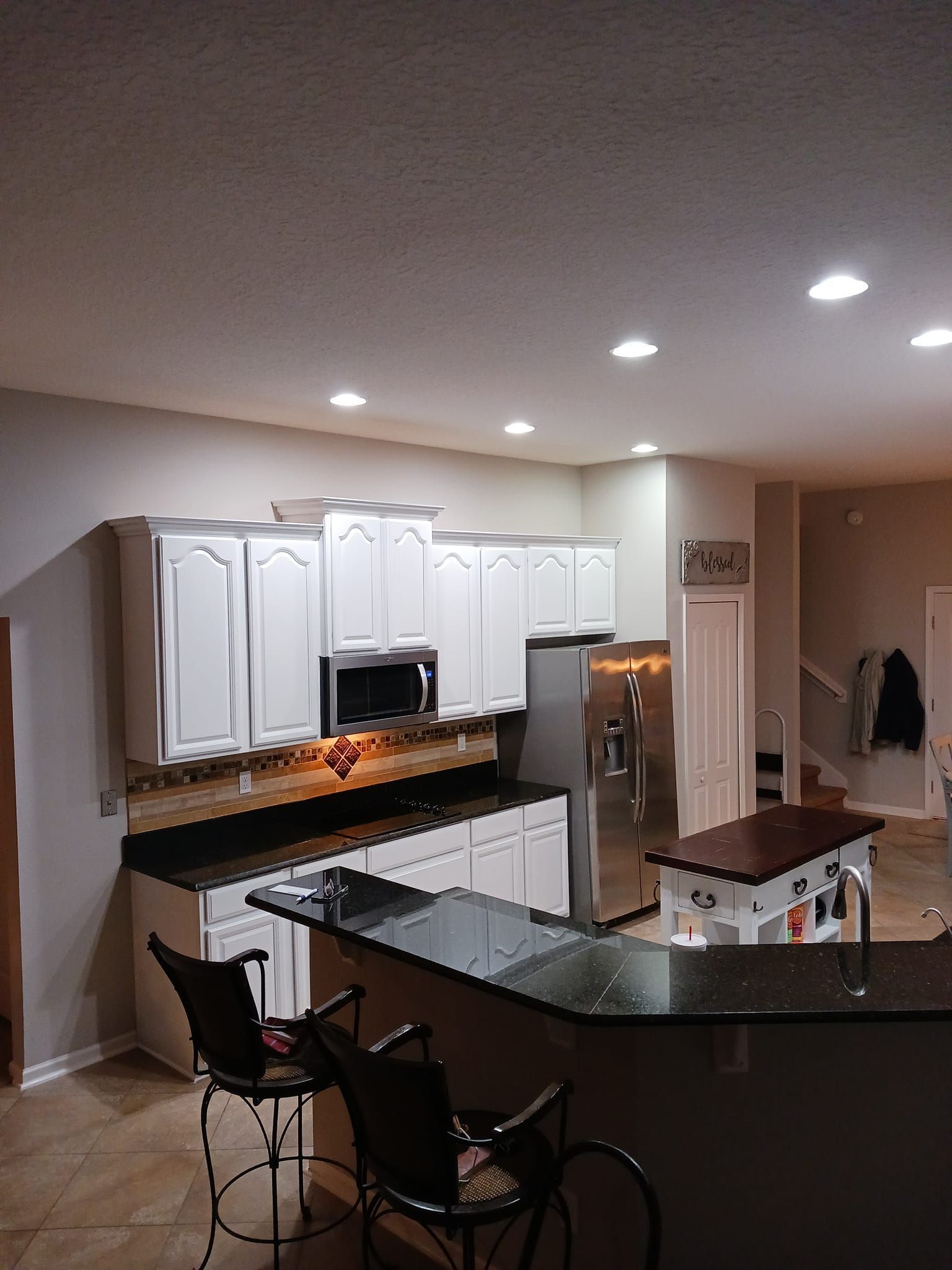 A kitchen with white cabinets, dark countertops, a stainless steel refrigerator, and two black bar stools.