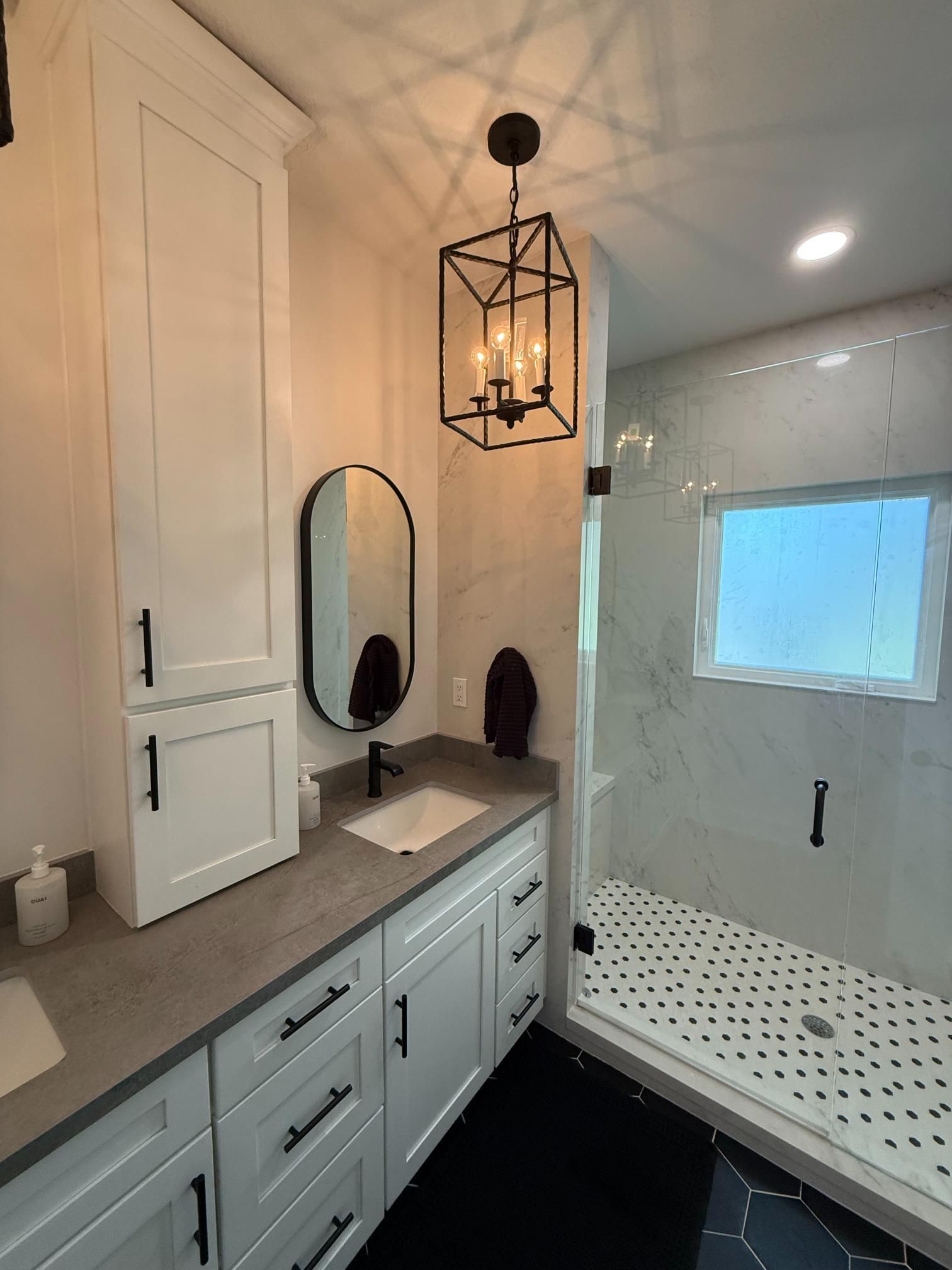 Bathroom with white cabinets, dark hardware, gray countertop, black framed mirror, shower, and pendant light.