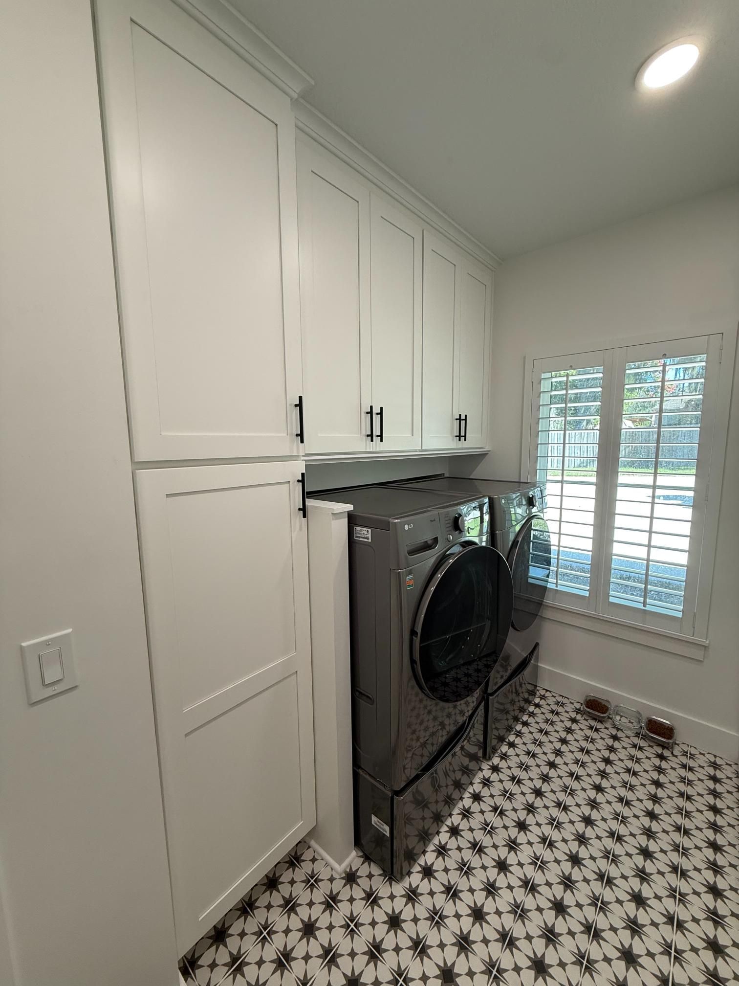 Laundry room with white cabinets, dark washer/dryer, patterned floor, and window with blinds.