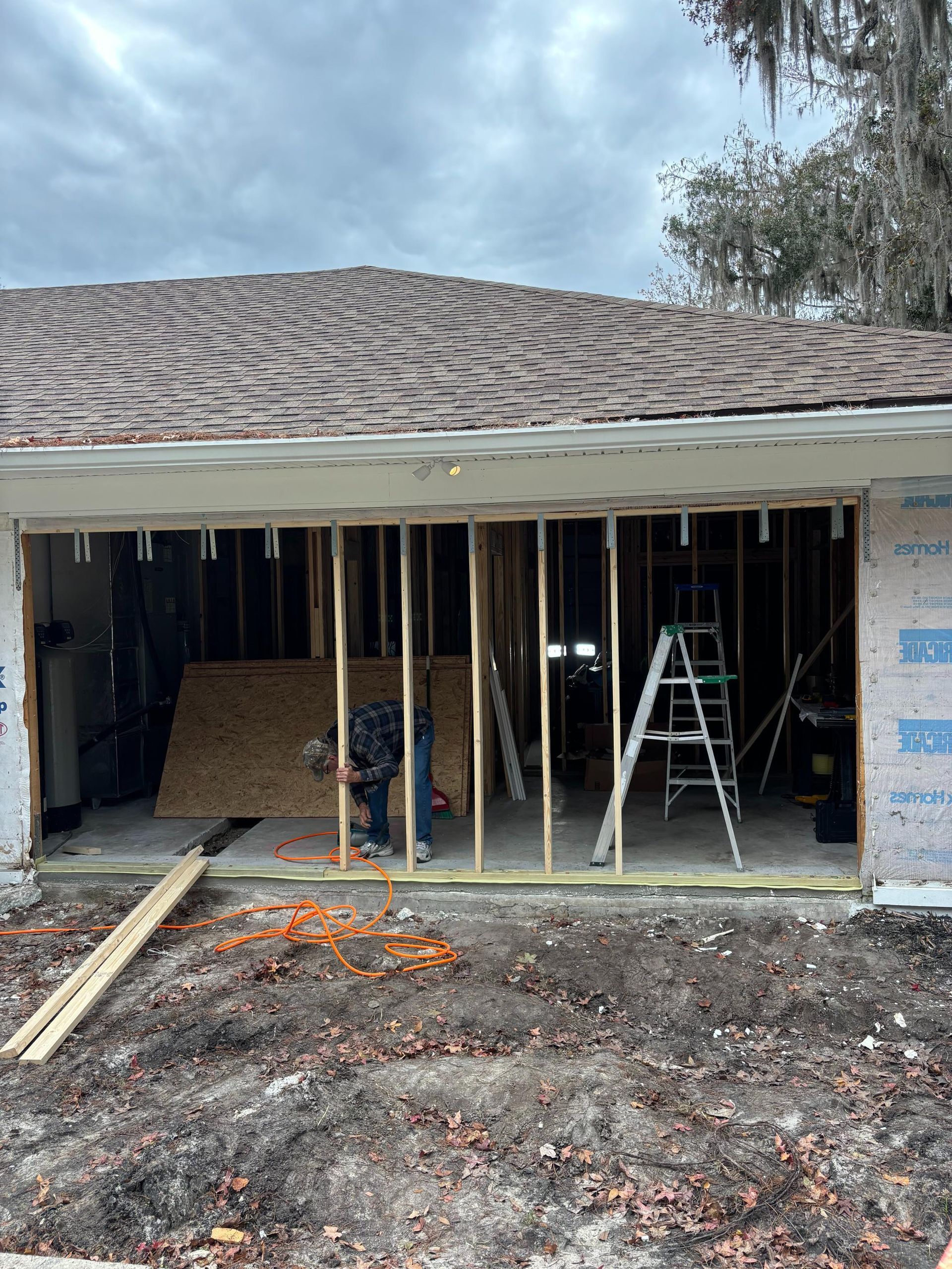 Garage under construction; a worker stands inside, framed by wooden studs; a ladder leans against a back wall.