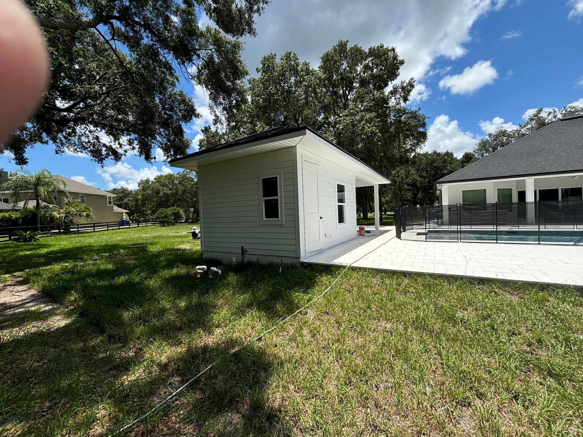 White shed in a grassy yard, next to a patio and pool, with trees and a blue sky in the background.