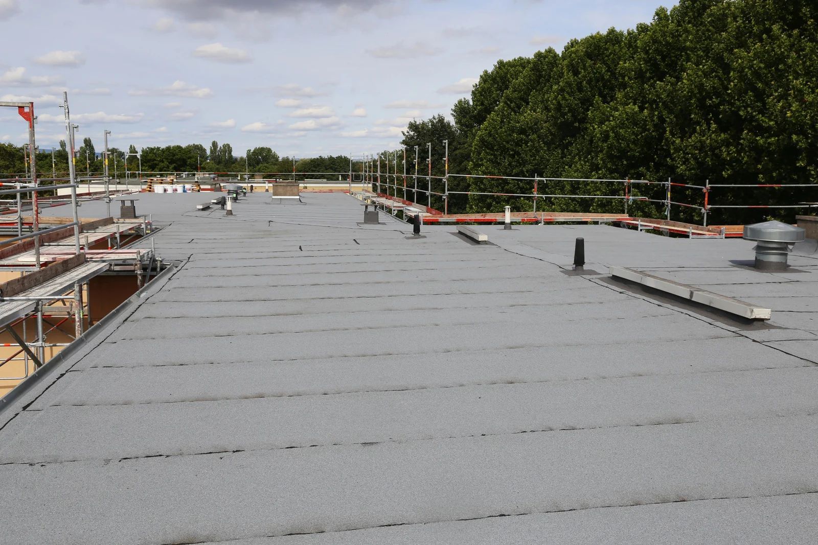 Flat gray commercial roof with various pipes and safety railings, with trees and a cloudy sky in the background.