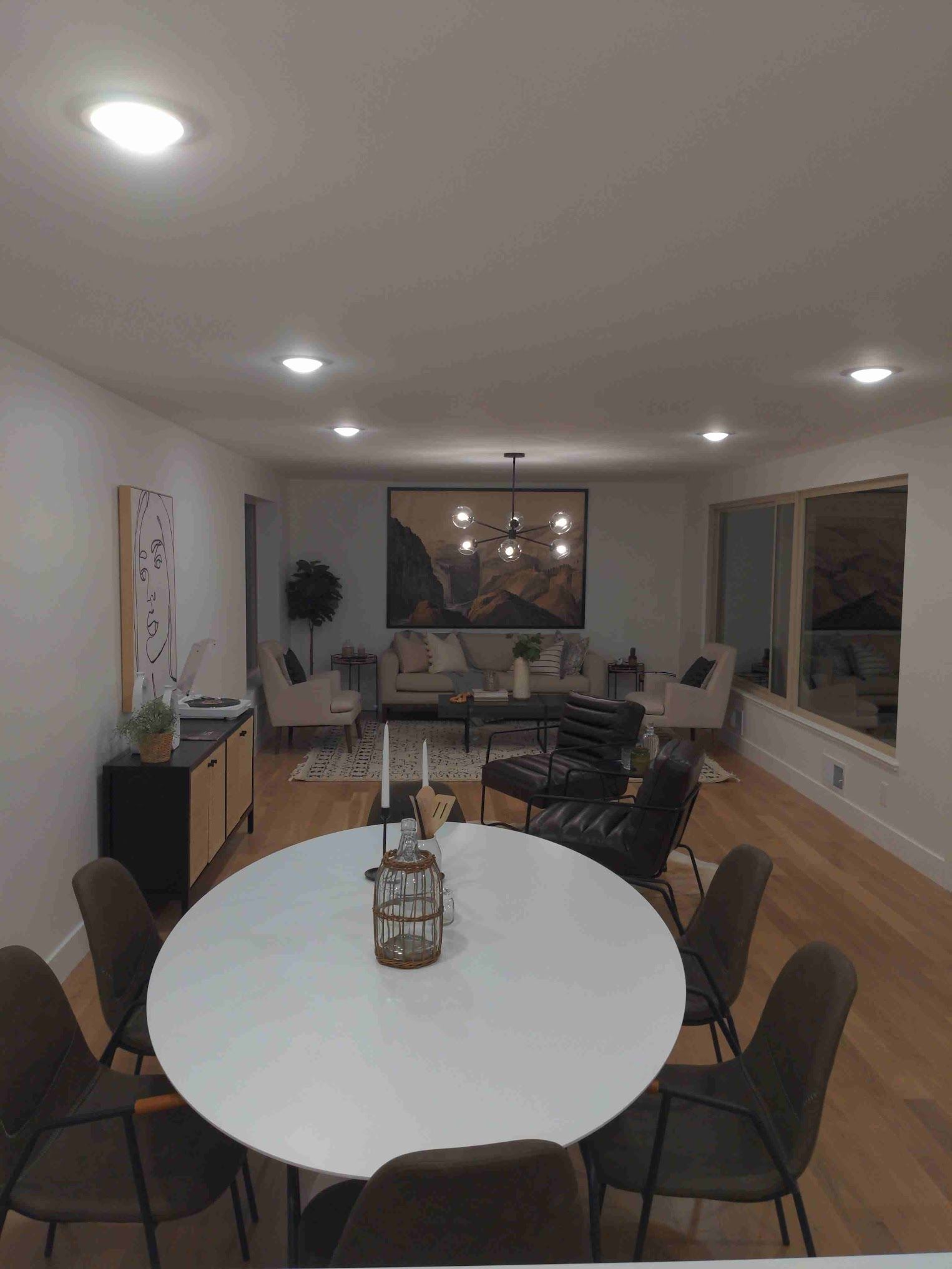 Spacious living room with dining table, chairs, sofa, and large artwork. White walls, wood floors, and recessed lighting.