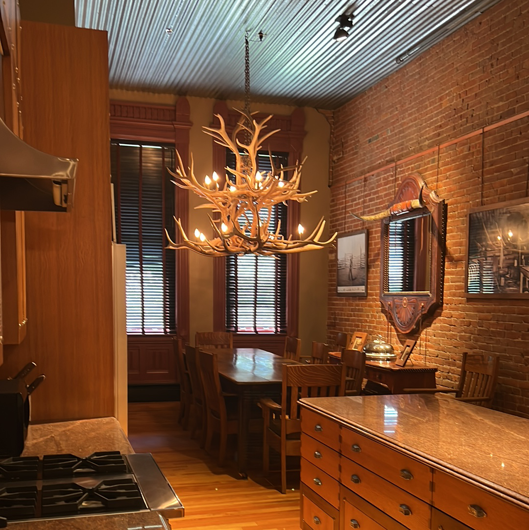 Rustic dining room with antler chandelier, exposed brick, and wooden furniture.