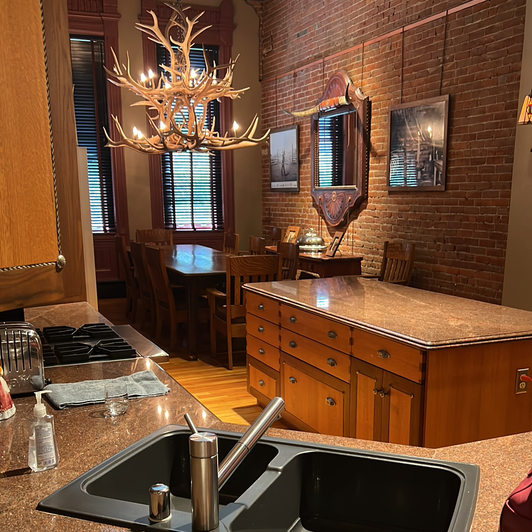 Kitchen view with antler chandelier, brick wall, wooden furniture, and a sink.
