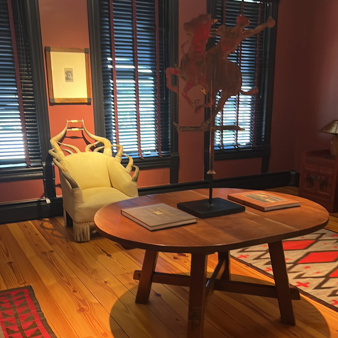 Room with table, books, chair, sculpture of a cowboy, windows with blinds, and red walls.