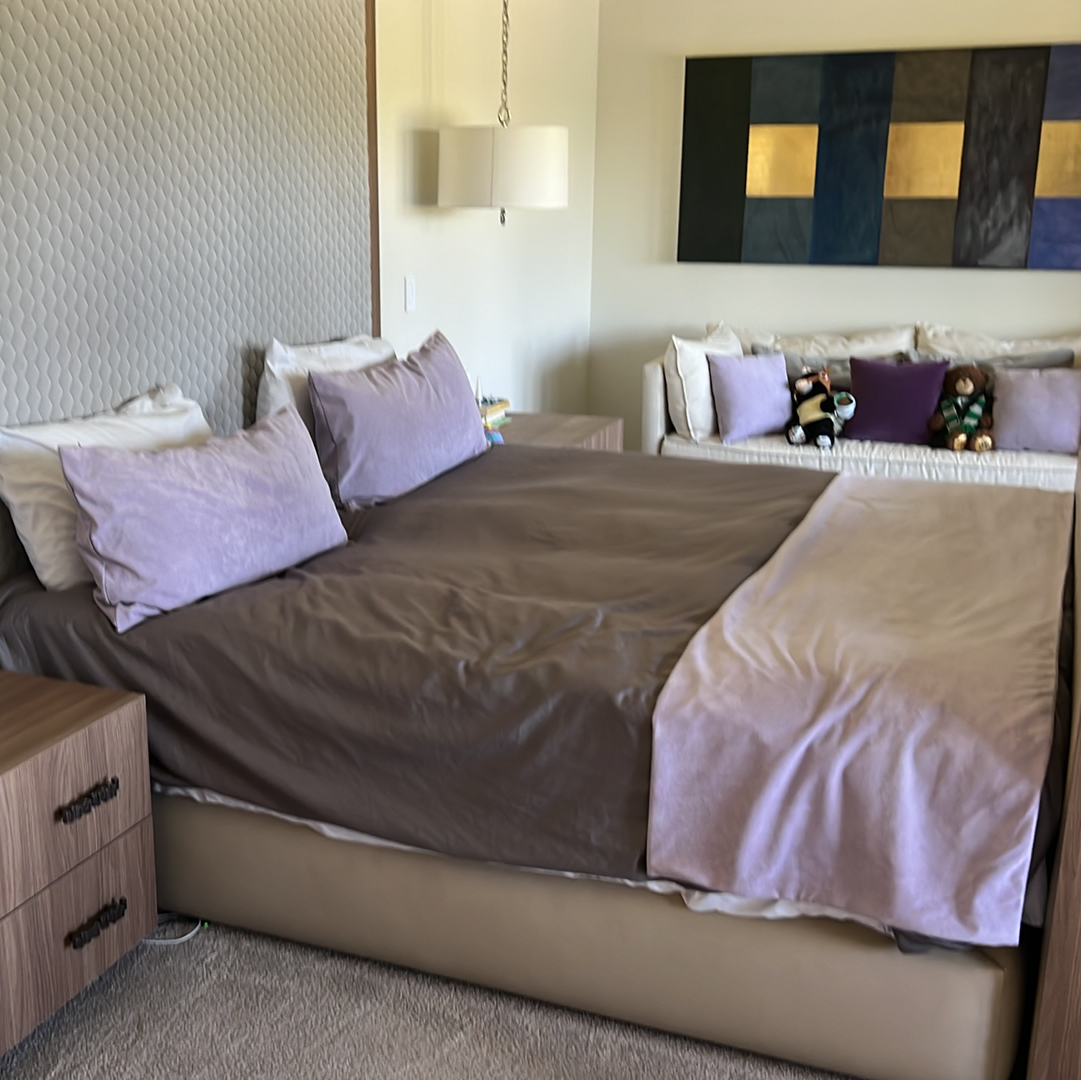 Bedroom with bed, nightstand, and small couch, soft purple and brown colors. Artwork on wall.