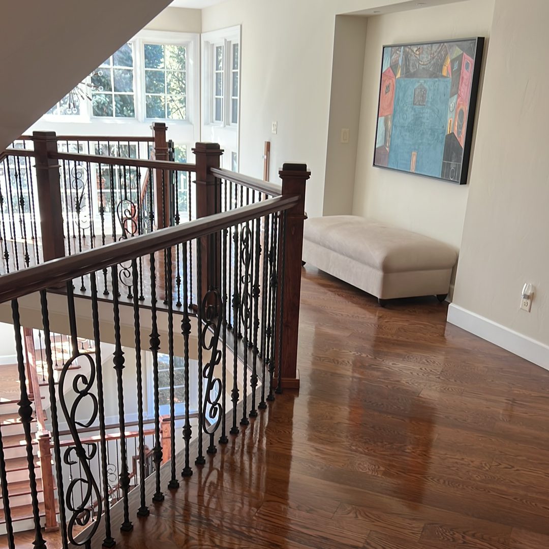 Hallway with staircase, wood floors, and art, cream-colored ottoman, neutral walls, and wrought iron railing.