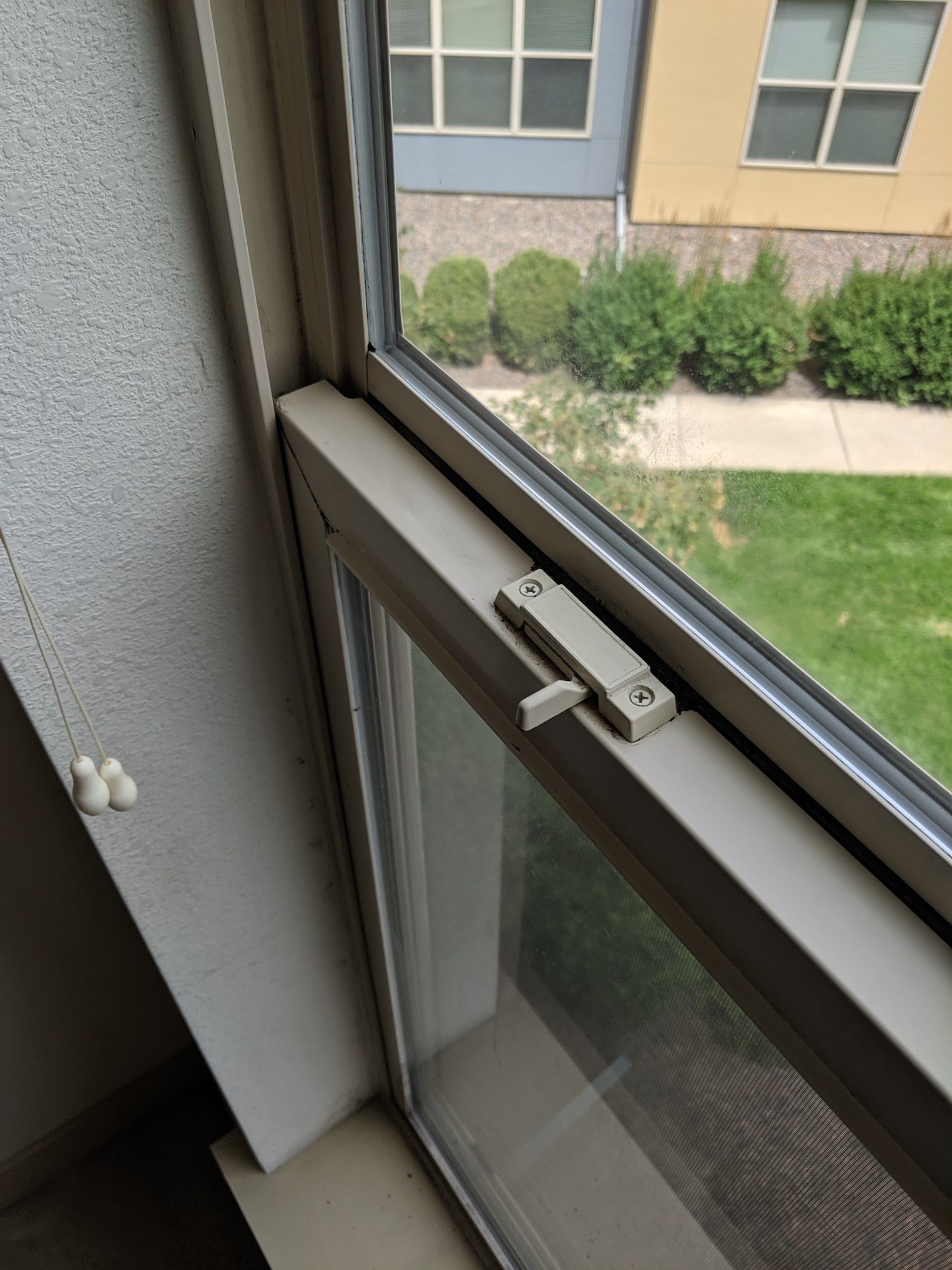 Beige window latch on a closed window overlooking a grassy yard and apartment buildings.