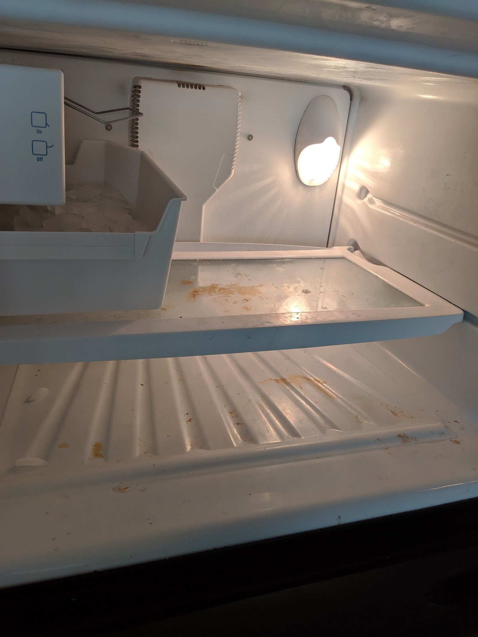 Empty mini-fridge interior with light on. White shelves and walls, with some food debris.