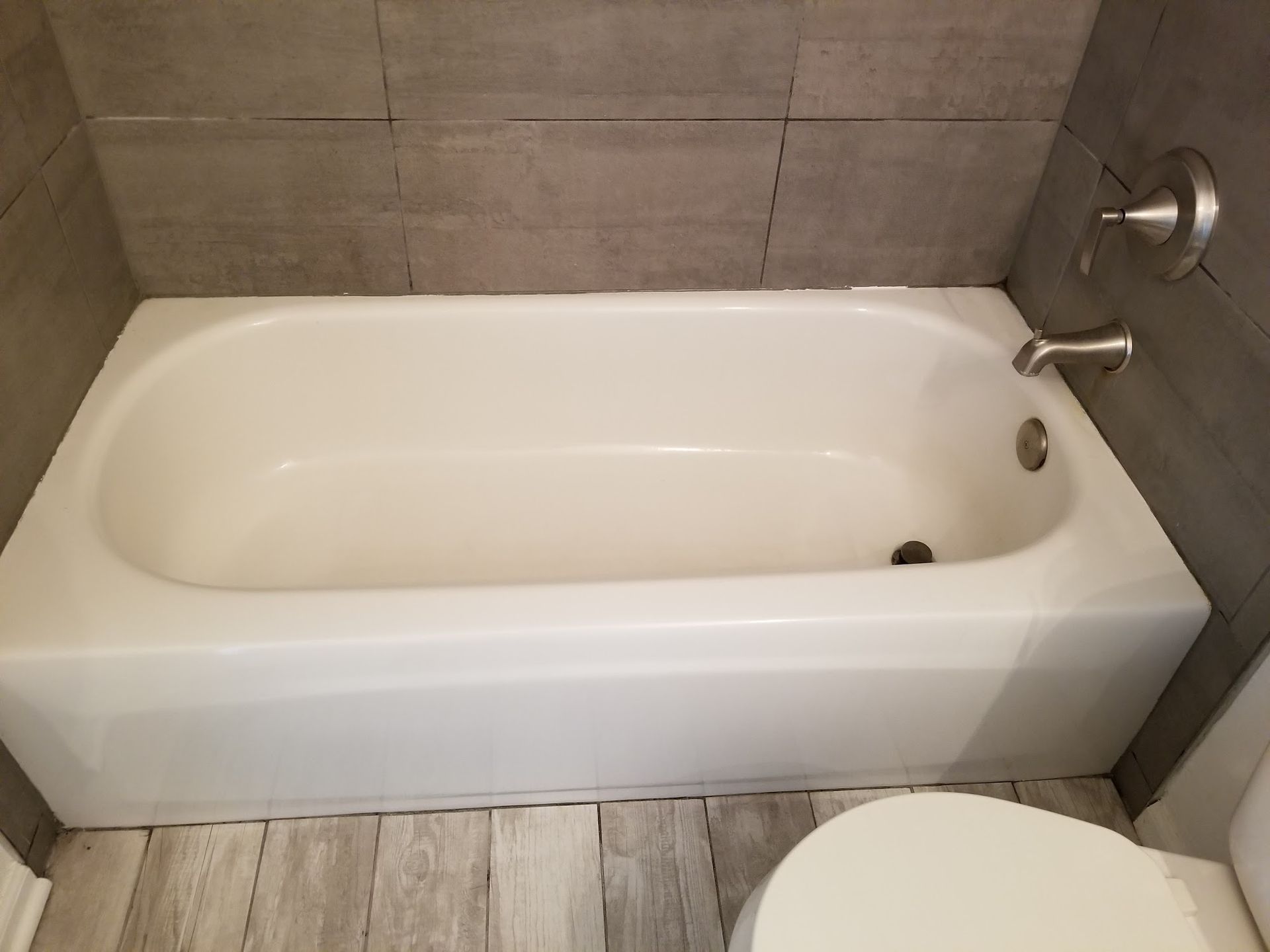 White bathtub against gray tile wall with toilet in the foreground.
